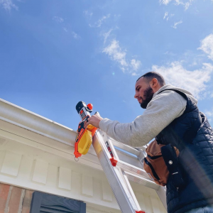 A man with a beard and a black vest is on a ladder securing it to a white gutter.