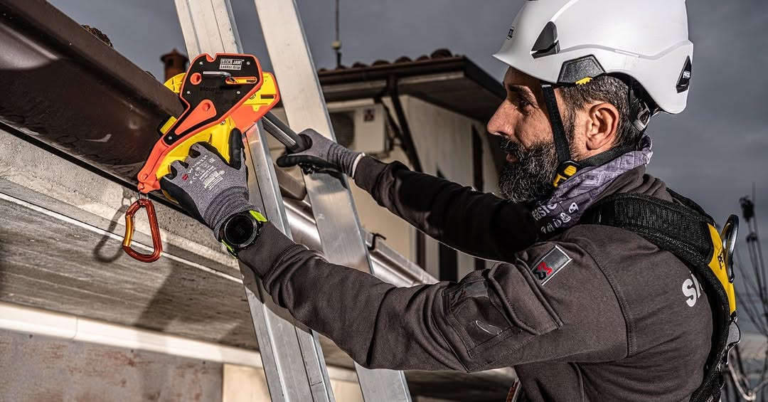 A person wearing a hard hat and safety harness attaches a Lock Jaw Ladder Grip to a ladder against a building.