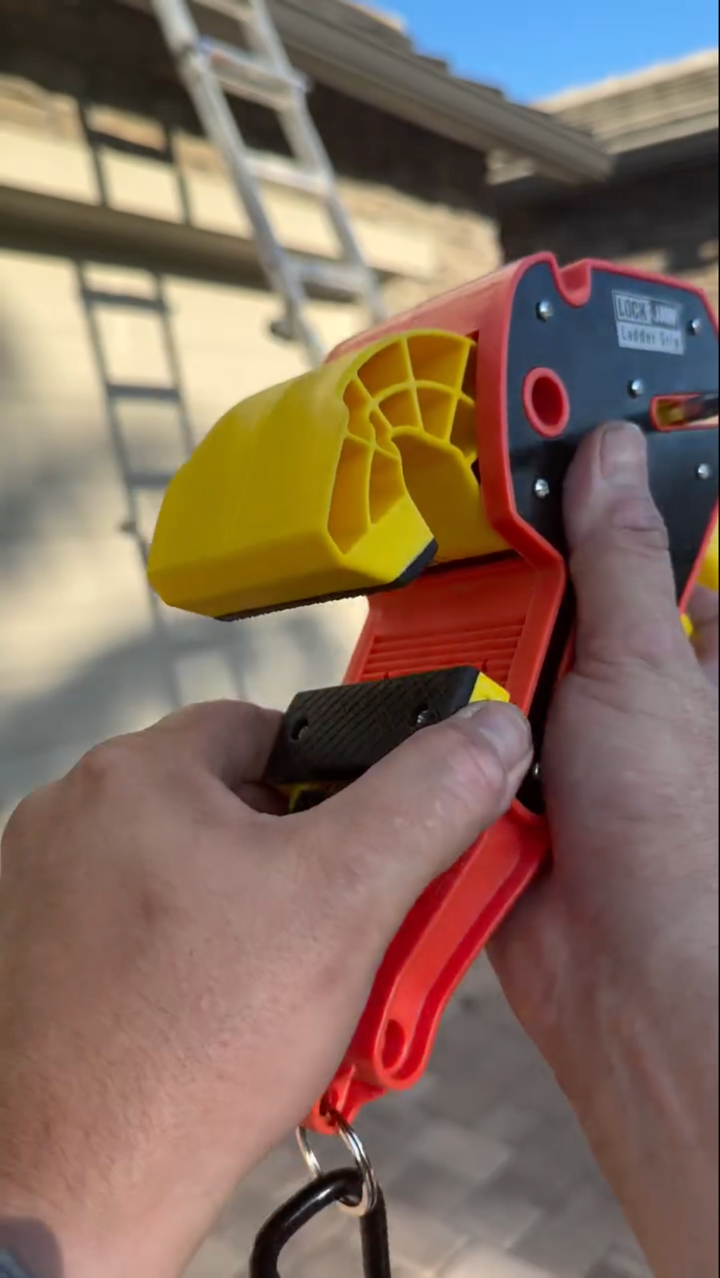 Hand held orange and yellow Lock Jaw Ladder Grip safety device being demonstrated by a person outdoors near a ladder.