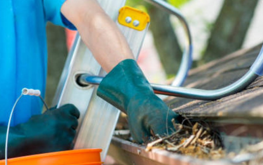 Gloved hands clearing packed leaves from a blocked gutter beside a ladder