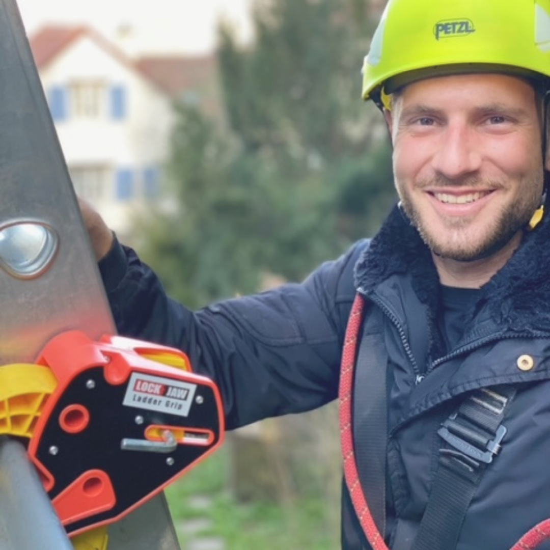 A smiling person wearing a yellow Petzl helmet and safety harness holds a Lock Jaw Ladder Grip attached to a ladder.