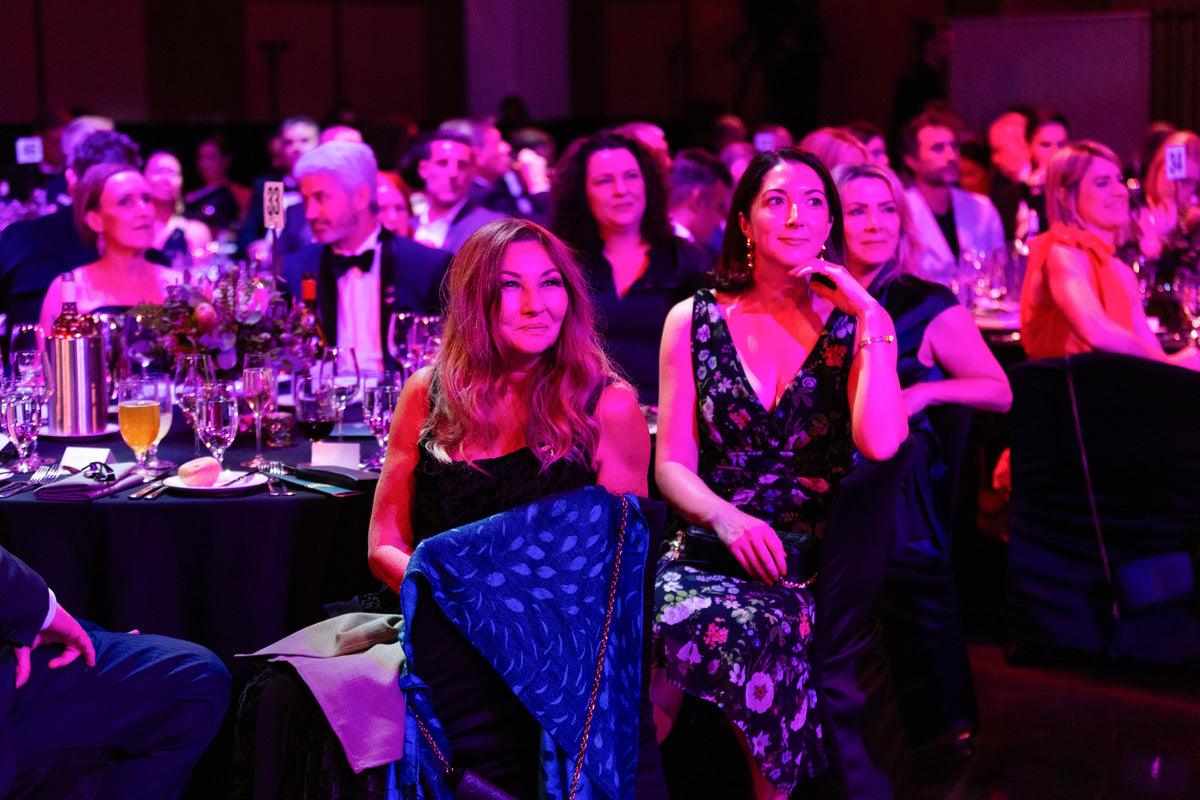Guests attend a gala dinner under purple lighting, with two women in evening dresses seated at a banquet table.