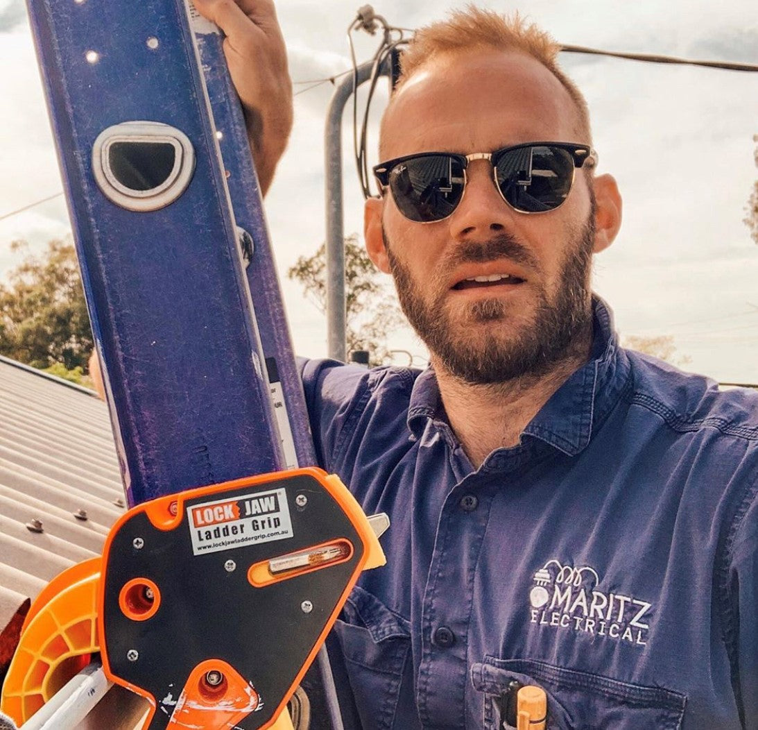 A fair-skinned man with short red hair, a beard, and sunglasses holds a ladder with a Lock Jaw Ladder Grip attached. He wears a blue "Maritz Electrical" shirt.