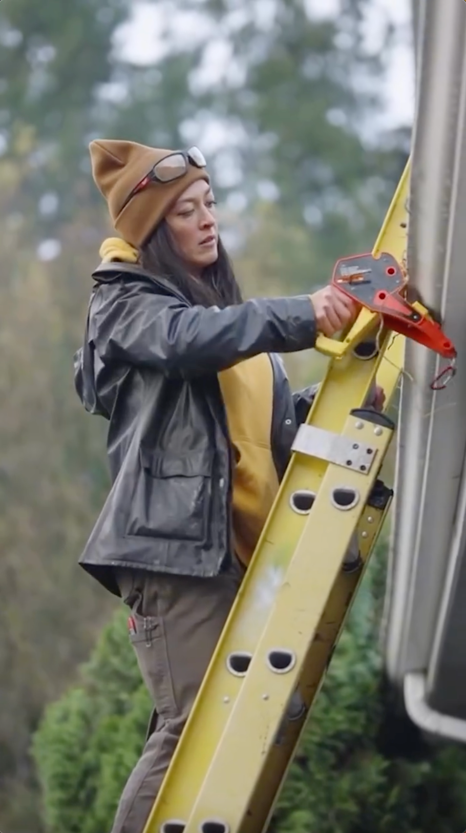 A woman with tan skin, wearing a brown beanie and goggles, adjusts a Lock Jaw Ladder Grip on a yellow ladder.