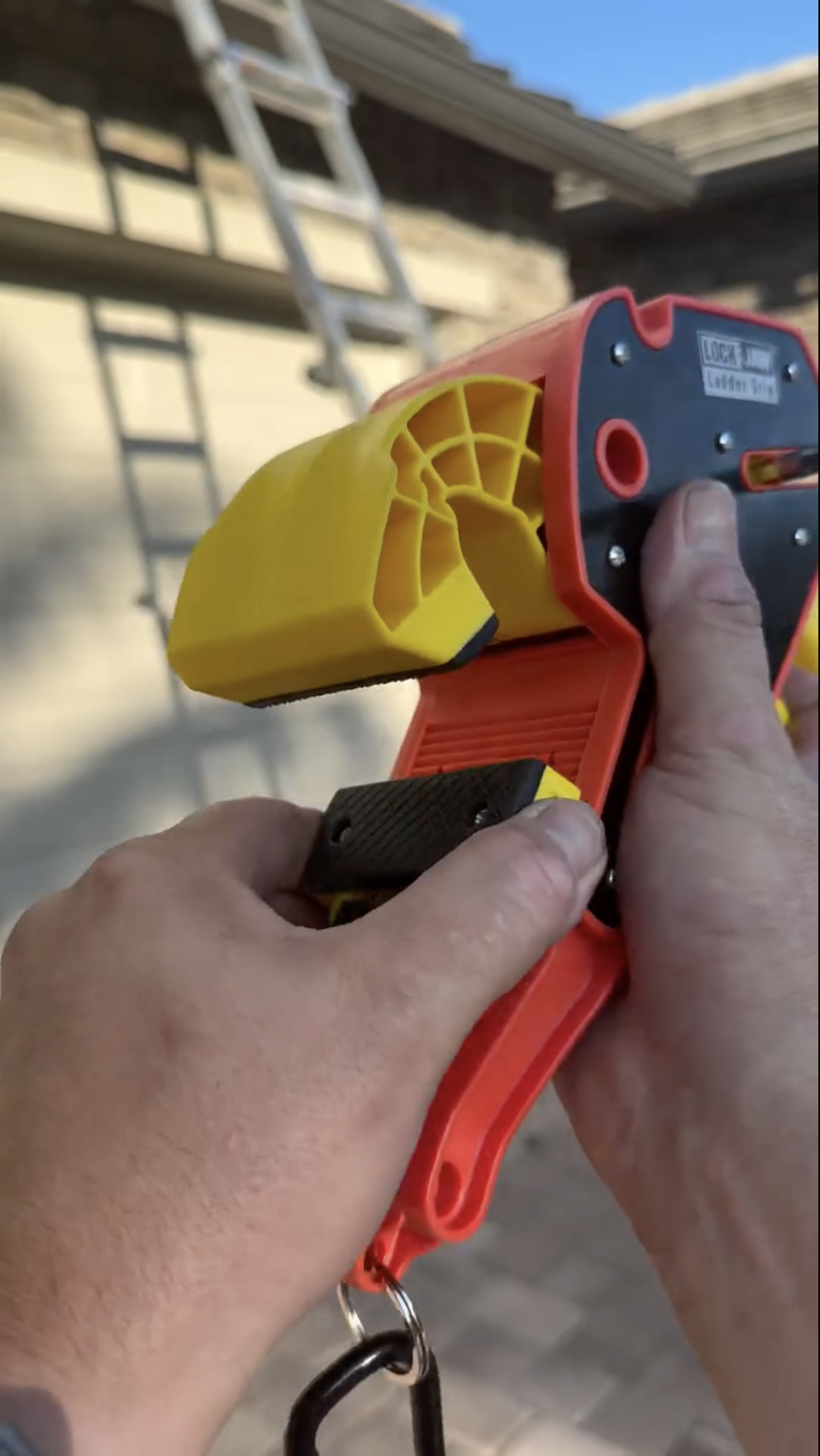 
Hand held orange and yellow Lock Jaw Ladder Grip safety device being demonstrated by a person outdoors near a ladder.