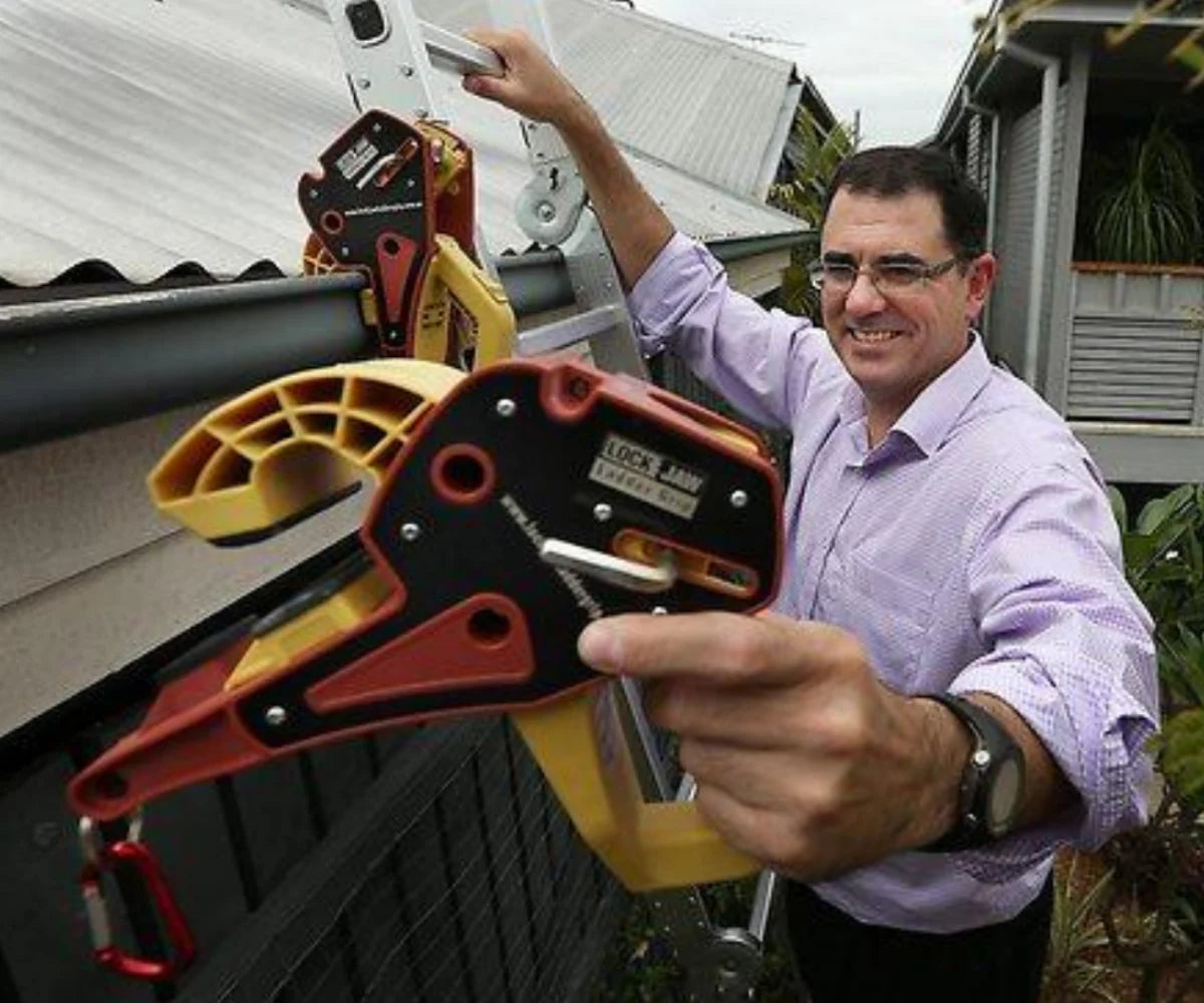 Man demonstrates Lock Jaw Ladder Grip on gutter while holding device toward camera beside extension ladder on roof.