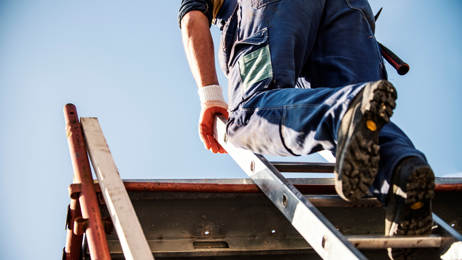 A person wearing work gloves and jeans climbs a ladder onto a roof under a clear blue sky.