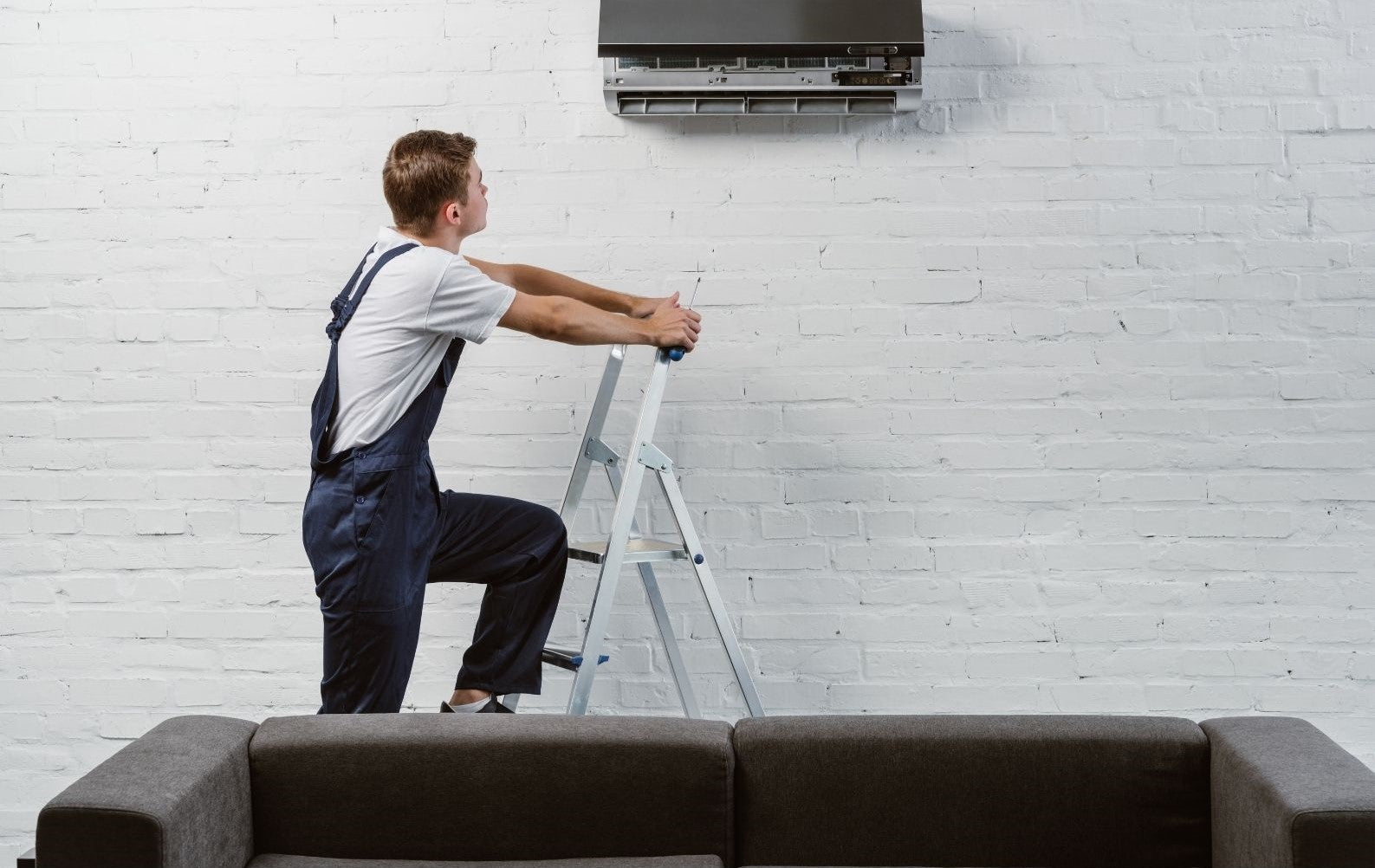 A person in overalls is climbing a walking step ladder to reach an air conditioner mounted on a white brick wall.