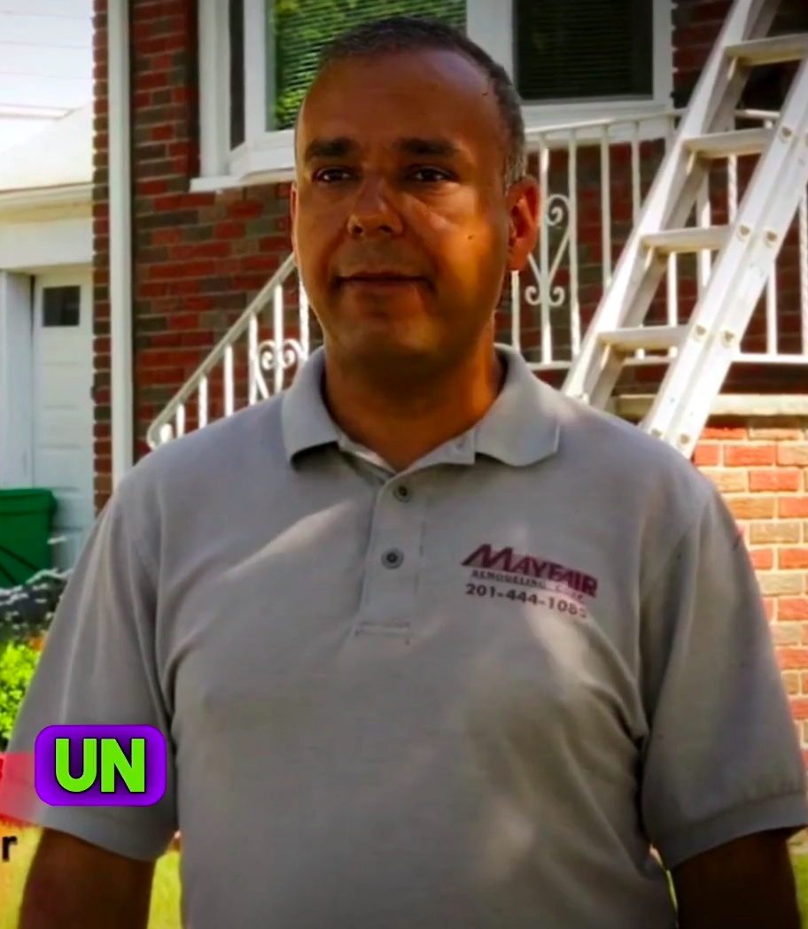 A man with short dark hair, appearing to be of Latin American descent, wearing a gray "Mayfair Aluminum" polo shirt, stands outdoors. A ladder and brick building are behind him.