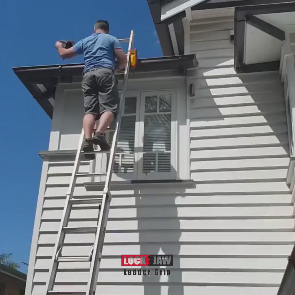 A person climbs down a ladder secured to the gutter of a white house with Lock Jaw Ladder Grips under a clear, blue sky.