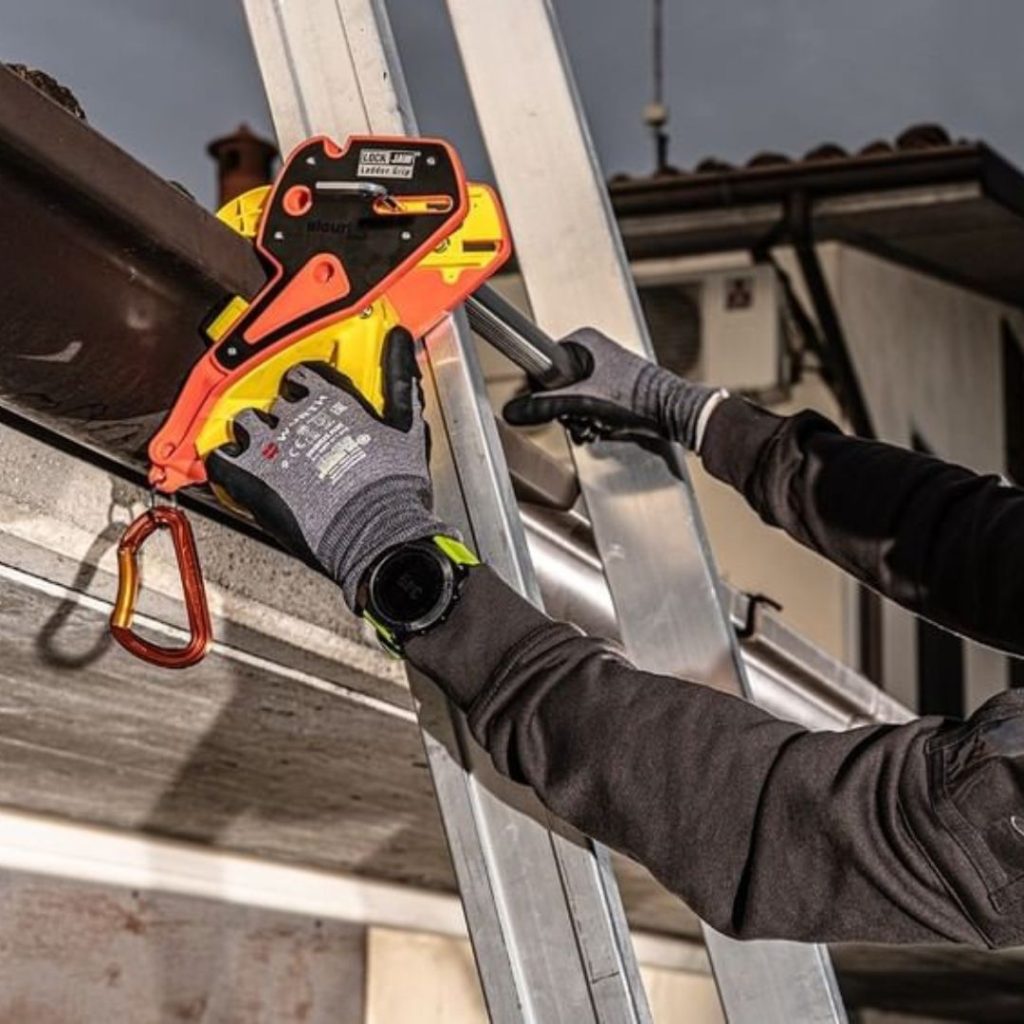 A person wearing gloves secures a Lock Jaw Ladder Grip to a ladder on a roof gutter.