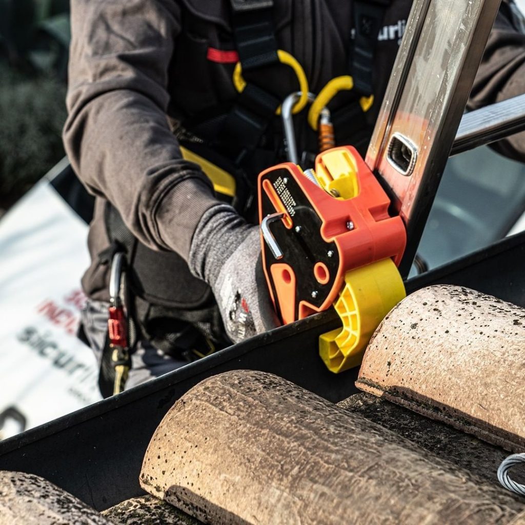 A person wearing a safety harness secures a Lock Jaw Ladder Grip to a roof. 