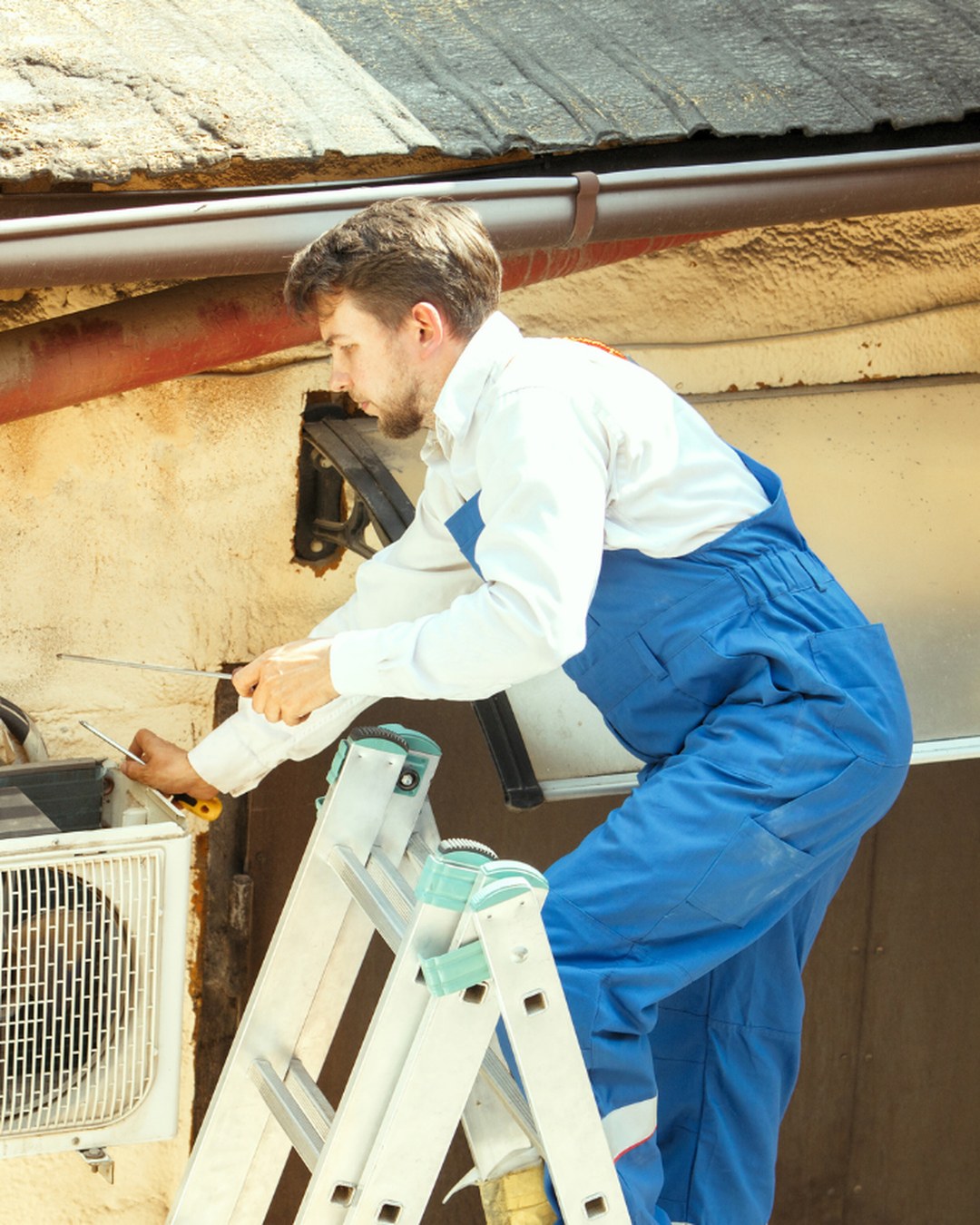 HVAC technician on an extension ladder servicing a wall-mounted condenser